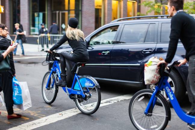 Woman and companion biking down West 4th Street on Citibike in traffic.